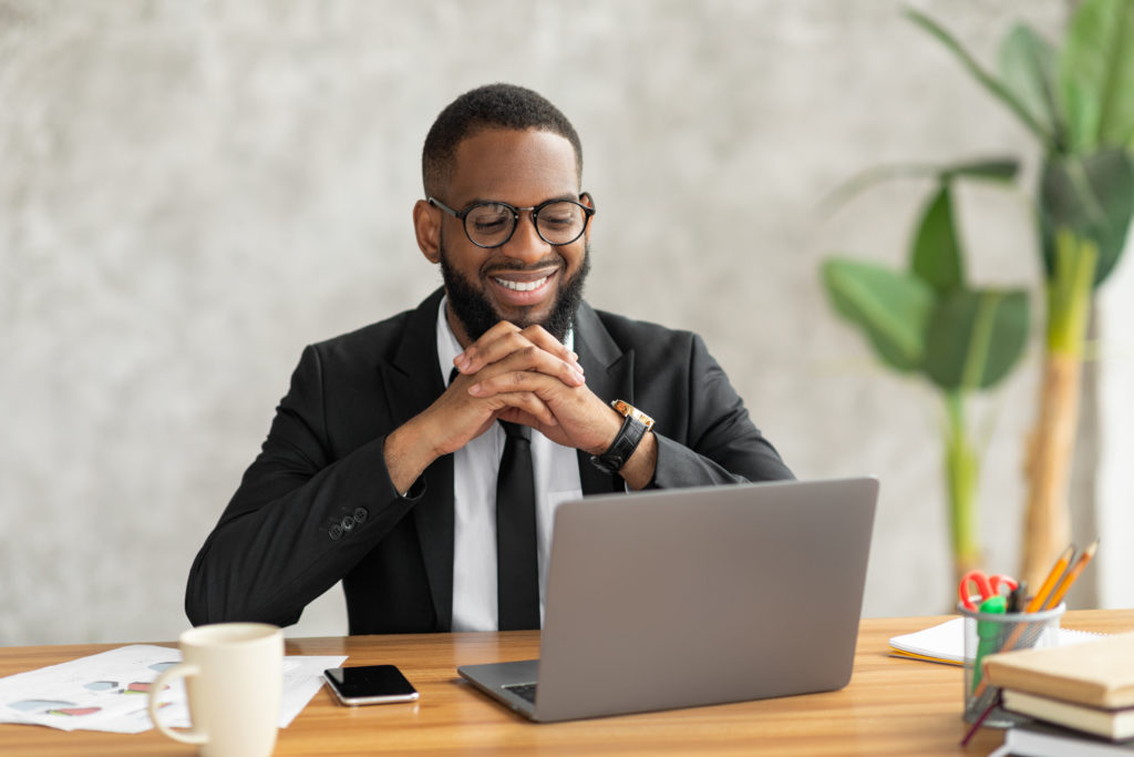 People And Technology Concept. Portrait of smiling young black man using laptop. Male worker in suit and eyeglasses sitting at desk, watching video, movie or webinar, taking break, making video call