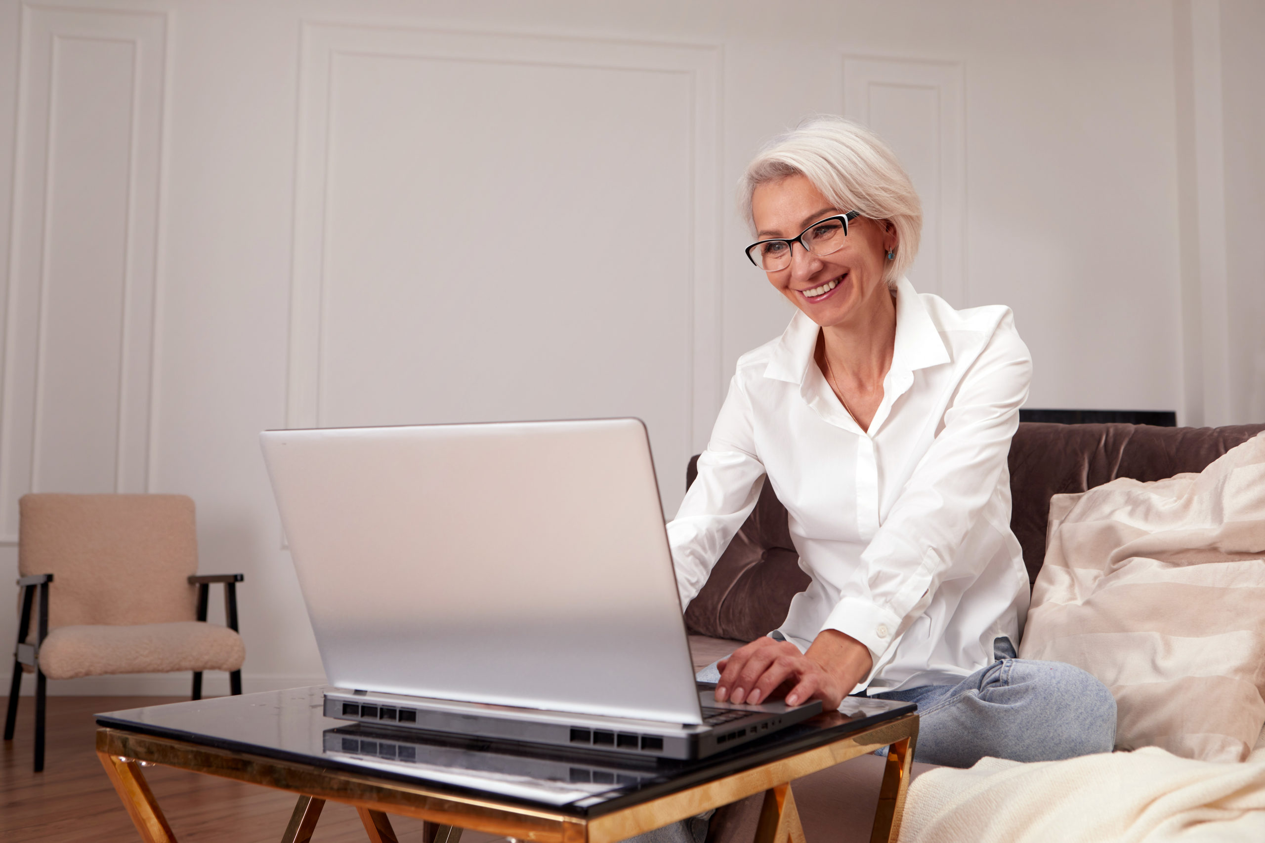 Happy 50 older mature middle aged adult woman holding laptop using computer sitting on couch at home. Smiling elegant senior grey-haired lady in glasses spending time with technology device livingroom